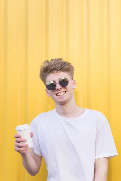 ガラスと白でスタイリッシュな若い男 シャツは 彼の手と笑顔でのコーヒーのカップから黄色の背景に立っています 一杯のコーヒーで幸せな若い男の肖像 — ストック写真