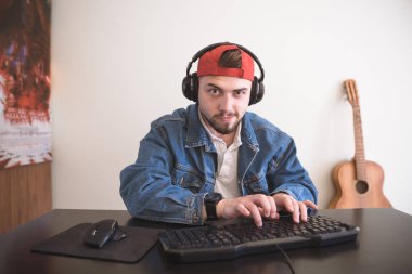 Funny man gamer plays computer games at home in a cozy room. Adult bearded gamer sits at the table and looks at the camera in a concentrated way. Video Game Concept. clipart