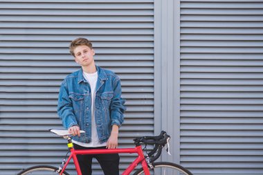 Bir bisiklet arka planda gri bir duvar ile bir kot ceket şık genç bisikletçi duruyor. Bir bisiklet ile şık bir adam portresi.