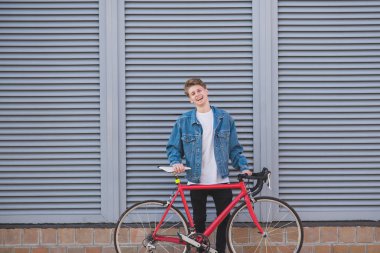 Şık, mutlu bir genç adam bir kot ceket ve gri bir duvar arka plan üzerinde kırmızı bir bisiklet portresi. Bir güzel, mutlu hipster adam duvar arka plan üzerinde bir bisiklet ile ayakta.