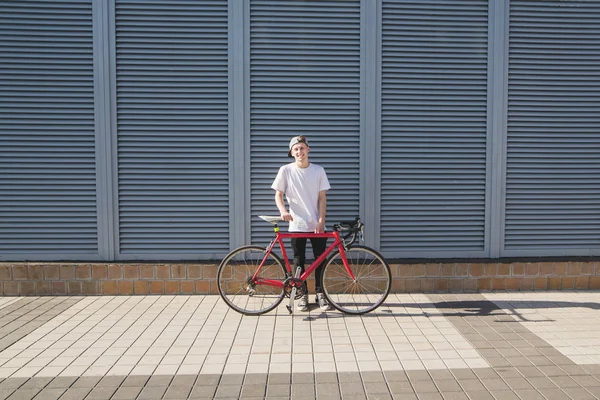 Student with a bicycle on the background of the wall. Happy young man in a white T-shirt and cap is standing on the background of a gray wall with a red bike and smiling.