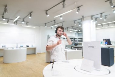 yakışıklı bir ışık teknik mağaza standları ve kulaklık müzik dinler. Alıcı becerikli alet elektronik mağazasında seçer. Seçme ve depodaki kulaklık satın alma
