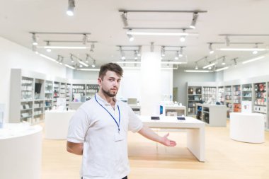 Girişte duruyor ve teknoloji dükkanına davet ediyor bir danışman portresi. Conlutant modern hafif elektronik mağazasından arka plan üzerinde duruyor