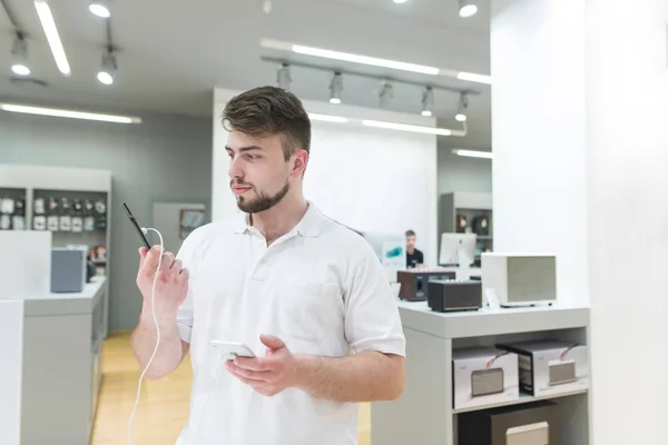 Buyer is in the electronics store in the mobile department with a smartphone in his hands. A handsome man chooses a smartphone in a modern technology store.