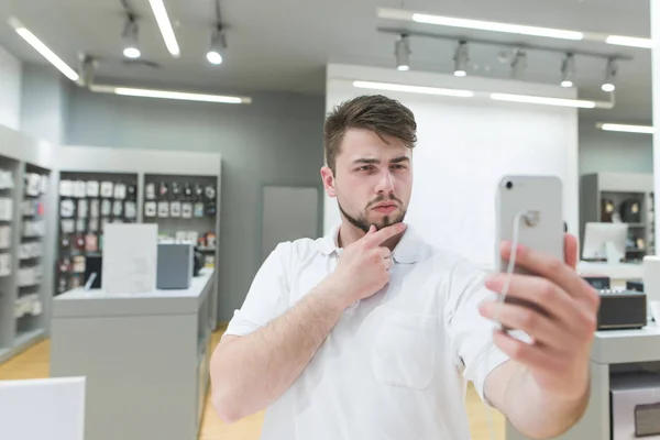 Funny man takes selfie on a smartphone in an electronics store. The buyer tests the front camera when choosing a smartphone. A man with a beard takes selfie in store technologies.