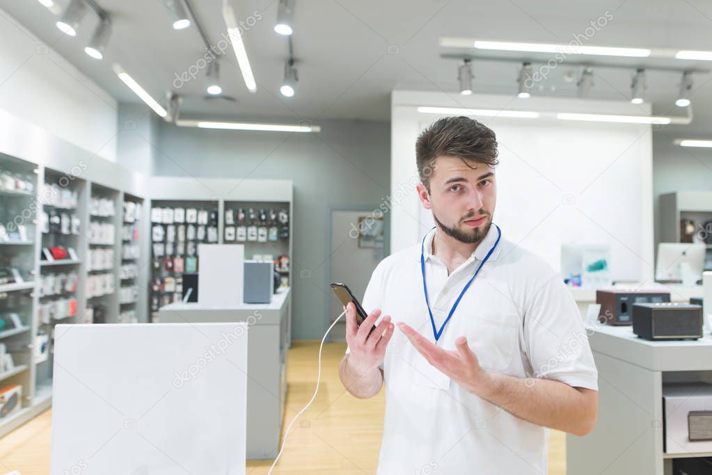 consultant is in the electronics store and recommends the smartphone in his hands. Portrait of a concert in a modern technology store. Work of the consultant in the electronics store.