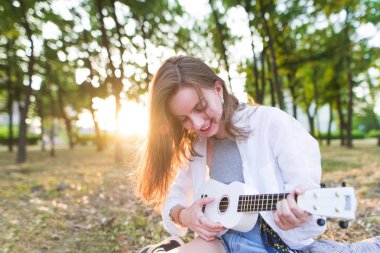 Mutlu gülümseyen kız çimenlerin üzerinde bir parkta oturur ve gün batımı arka planı beyaz bir ukulele üzerinde çalış. Şık bir kız parkta bir müzik aleti çalıyor.