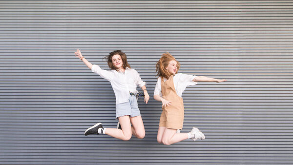 Two stylish happy girls jump together on a gray background. Portrait of smiling fashionable girlfriends in motion that jump on a dark background.