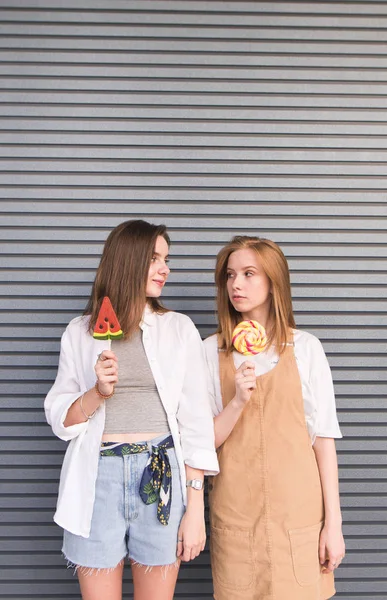 きれいでスタイリッシュな女の子はお菓子を手に立っている 灰色の壁の背景にお互いを見てします ロリポップと魅力的な女の子の肖像画 — ストック写真