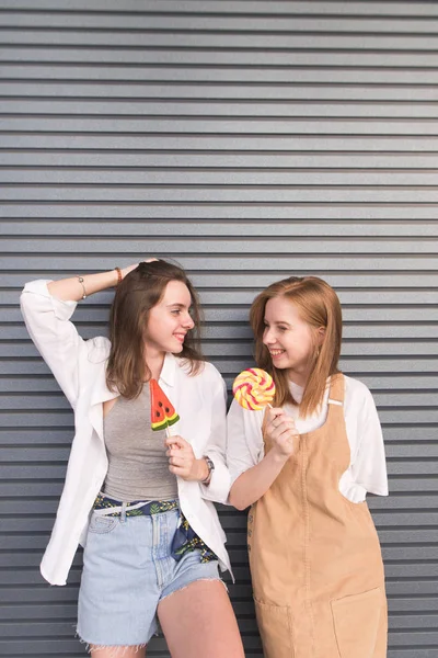 Gri bir karşı ellerinde şeker ile iki gülümseyen şık kız arkadaşım arka plan duvar. Lolipop ellerinde bir gri zemin üzerine iki mutlu kız şık hafif giysiler koymak.