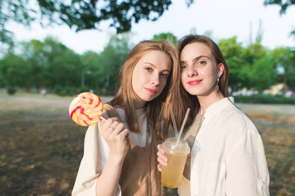 两个有魅力的女朋友 手里拿着棒糖和柠檬水 摆在公园里拍照 两个微笑 时尚的女孩的肖像站在公园里 看着照相机 — 图库照片