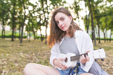 Ukulele elleri ile bir parkta oturan ve kameraya bakarak çekici bir kız portresi. Pretty genç kız parkta ukulele çalış.
