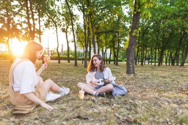 Two happy friends are sitting in the park and having a rest. One plays on a ukulele another eating lollipop. Recreation with a girlfriend in the park at sunset.