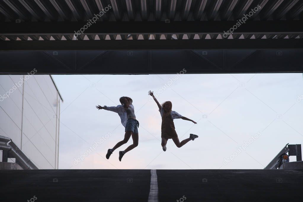 Silhouette of two young girls who jump on. Silhouette of two friends in a jump on the background of the sky near modern architecture.