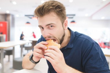 Yakışıklı bir fast food restoran oturur, bir hamburger yiyor ve kameraya benziyor. Öğle Yemeği kötü bir lokanta var. Fastfood kavramı. öğrenci hamburger bir fast food restoran yiyor..