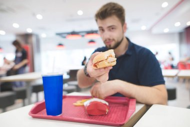 Adam bir lokanta tepsi ile parlak bir restoranda bir masada oturuyor ve adamcağız hamburger kamerada gösterir. İçinde bir adam elinde bir hüner iştah açıcı burger.