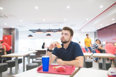 Öğrenci sakallı bir fast-food restoran lokanta tepsisinde onun elinde hamburger ile ile masada oturur ve yiyor. Adam yemekleri lezzetli lokanta ile. Fast food lezzetli kavramı.