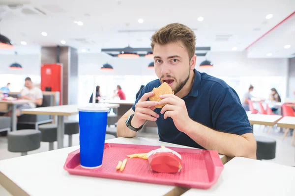 Man met een baard zit aan een tafel met een lade in een fast-food restaurant met een hamburger in zijn handen en kijkt naar de camera. Student is het eten van fast-food bij het fast-food restaurant. — Stockfoto