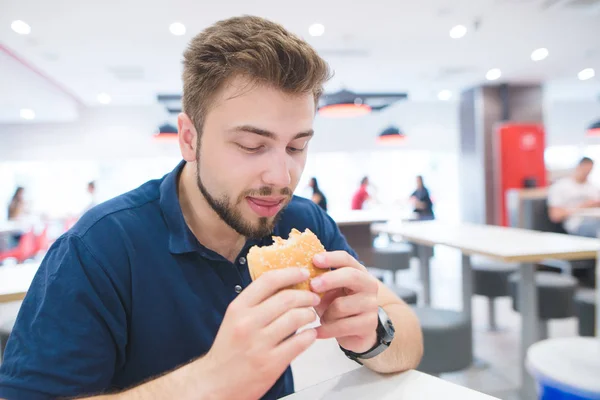 明るいファーストフードのレストランに座っていると食欲を彼の手でハンバーガーを見てひげを持つ男の肖像画 ファーストフードのレストランの手でハンバーガー学生 — ストック写真