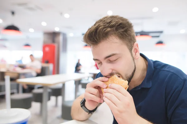 Homem com prazer morde um hambúrguer apetitoso no fundo de um restaurante de fast food. O estudante come fast food no restaurante. Conceito de fast food . — Fotografia de Stock