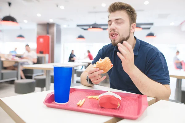 O homem está sentado em uma mesa em um restaurante de fast-food e come um menu de hambúrguer, uma bebida e batatas fritas. O homem está gostando de fast food. Conceito de fast food . — Fotografia de Stock