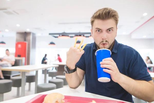 Grappige mens met frietjes en een glas van koel drankje zit in een restaurant en kijkt naar de camera. Emotionele man drinkt en eet van Franse frietjes in een restaurant. — Stockfoto