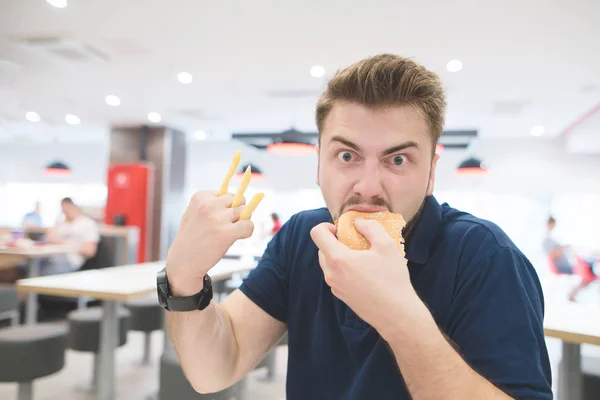 Портрет смешного эмоционального человека с картошкой фри вместо пальцев, который ест бургер в ресторане быстрого питания и смотрит в камеру. Концепция быстрого питания — стоковое фото
