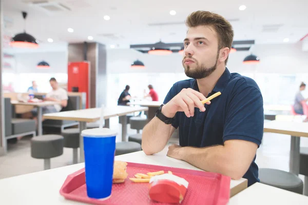 Retrato de um homem com batatas fritas em seus braços sentado em um restaurante de fast food brilhante e comer fast food. O estudante senta-se a uma mesa com uma bandeja na qual uma bebida fresca, um hambúrguer e batatas fritas . — Fotografia de Stock