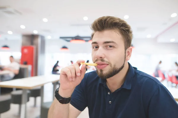 El hombre come papas fritas en un restaurante de comida rápida y mira a la cámara. Retrato de un estudiante con barba sentado en un restaurante con papas fritas en las manos y mirando a la cámara. Comida rápida . — Foto de Stock