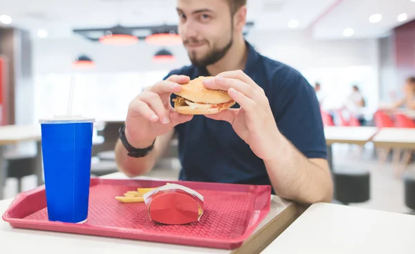 Étudiant avec un hamburger appétissant et plateau de restauration rapide se trouve à la table de restauration rapide. L'homme mange de délicieux aliments nocifs. Concentrez-vous sur le burger appétissant dans les mains d'un homme . — Photo