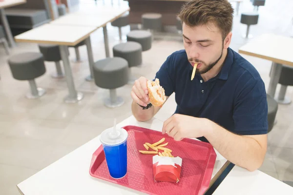 Yetişkin adam sakalı ve bir siyah t-shirt masada oturur ve fast food hamburger, yiyor patates kızartması ve bir fast food restoranında içecekler. Öğrenci, patates, Hamburger yiyor bir fast food restoran. — Stok fotoğraf