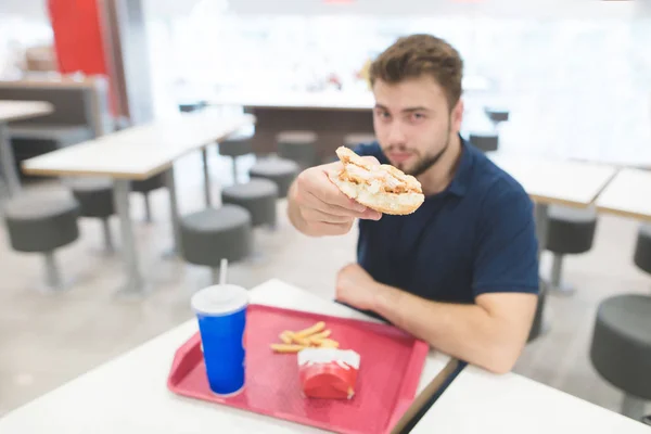 Ein Mann sitzt an einem Tisch in einem Restaurant mit einem Tablett voller Speisen und Getränke und zeigt einen Buggy-Burger in die Kamera. Ein Student isst im Restaurant leckeres Essen. Fokus auf den Burger. — Stockfoto
