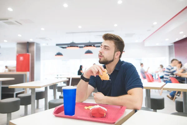 Retrato de un hombre guapo con barba sentado en una mesa en un restaurante lleno de luz con una bandeja de comida y hamburguesa en las manos. Estudiante come una hamburguesa en un restaurante de comida rápida. Comida rápida . — Foto de Stock