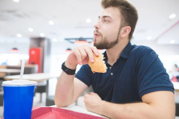 Közelkép portré egy ember, egy burger, a keze alatt, hogy ebéd egy gyorsétteremben. Diák eszik, szendvicset étterem háttérrel. Fast Food koncepció. Összpontosítani a burger. — Stock Fotó