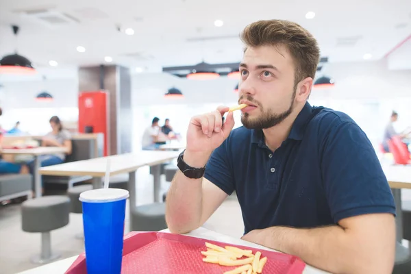 Портрет человека, который ест картошку фри в легком ресторане быстрого питания и смотрит в сторону. Студент с бородой и черной футболкой ест фаст-фуд из жевательного подноса. Концепция быстрого питания . — стоковое фото