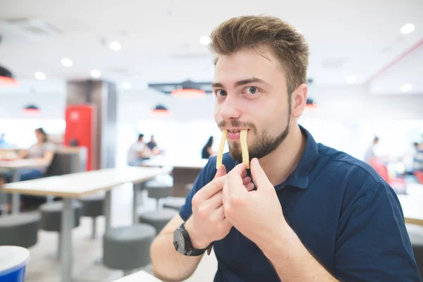 Zabawny człowiek z brodą wstawiane frytki zamiast jego zęby i patrzy w kamerę. Zabawna osoba jeść frytki w restauracji fast food. Grając z jedzeniem. — Zdjęcie stockowe