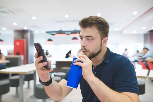 Der Student trinkt ein kühles Getränk aus einem blauen Glas und bedient sich eines Smartphones vor dem Hintergrund eines Fast-Food-Restaurants. Mann isst Fast Food, trinkt ein Getränk und blickt auf Smartphone. — Stockfoto