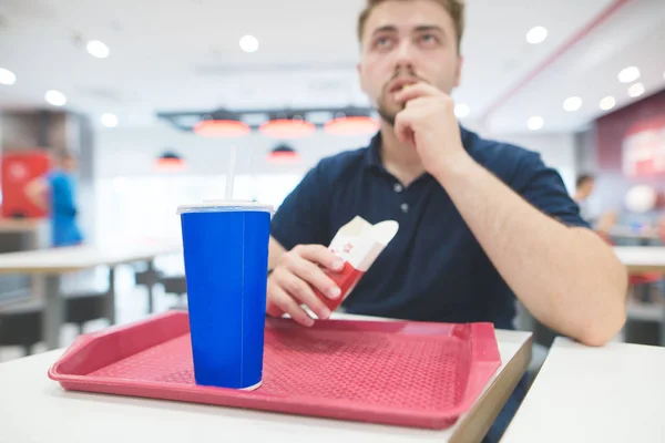 Adam masada restoranda kırmızı tepsisinden hızlı yemek yiyor. Serin tatlı içki ve arka plan üzerinde mavi bir bardak patates kızartması yiyen bir adam. Cam üzerine odaklanır. Fast Food kavramı. — Stok fotoğraf