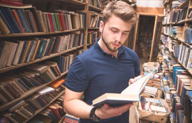 Bir sakallı yakışıklı adam elinde bir kitapla eski kütüphanede standları ve okuyun. Bir öğrenci bir kitapla kütüphanede portresi. Edebiyat kütüphanede arama.