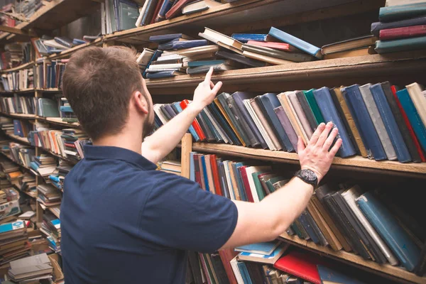Man chooses a book from the shelves of an old library. A student is looking for books in a library. The choice of literature. Shelves with books and a man\'s back in a blue t-shirt.