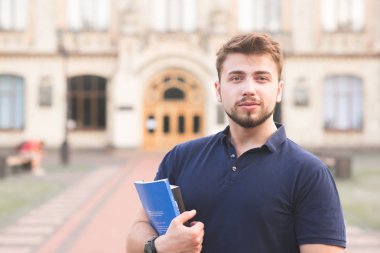 Kitaplar ve ellerini dizüstü bilgisayarlar ile ayakta ve kameraya bakarak bir güzel öğrenci adam portresi. Sakalı ve Üniversite binası arka planı elinde kitap olan adam