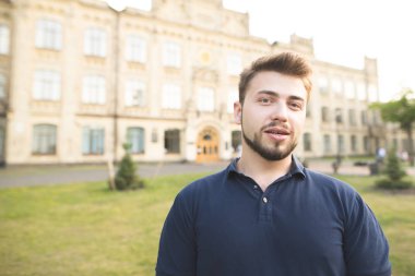 Arka planda üniversite kampüs binalarda bir sakal konumu olan yakışıklı bir adam portresi. Kamera ve gülümsüyor mutlu öğrenci siyah T-shirt olarak görünüyor.