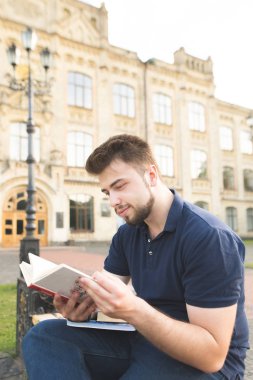 Gülümseyen öğrenci kampüsünde bir bankta bir bankta oturur ve bir kitap okur. Üniversite binasının arka plan açık havada üzerine bir kitap okuma sakallı gülümseyen bir adam portresi