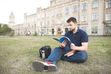 Üniversitesi Kampüsü çimenlerin üzerinde oturan ve bir kitap okuma bir öğrenci portresi. Adam Üniversite binası arka planda açık bir sakal çalışmalarla.