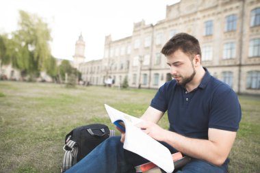 Konsantre öğrenci üniversite Binası yakınındaki çimenlerin üzerinde oturan bir kitap okur. Üniversitesi Kampüsü çimenlerin üzerinde oturan bir kitap okuma bir sakallı bir adam portresi.