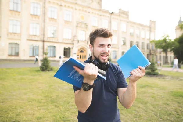 Defter ve kitaplar onun elinde ile üniversite binasının arka plan üzerinde duran ve kamera seyir mutlu bir öğrenci portresi. Neşeli sakallı adam ve kitap bir kampüs elinde