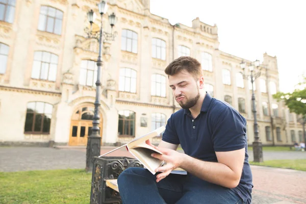 Kitaplar ile bir üniversite kampüsünde bir bankta oturmuş ve eğitim sinirli bir öğrenci portresi. Üzgün adam sakalı ve Kitaplar üniversite binasının arka planı.