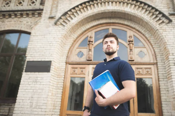 Portrait of a beautiful man with a beard holding books in his hands against the background of an old building. Student on the background of the door and building of the university.