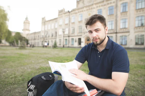 Yakışıklı adam elinde bir kitapla bina Üniversitesi arka planı çimenlerin üzerinde oturur ve kamera görünüyor. Elinde bir not defteri ile bir çim kampüste oturan bir öğrenci portresi