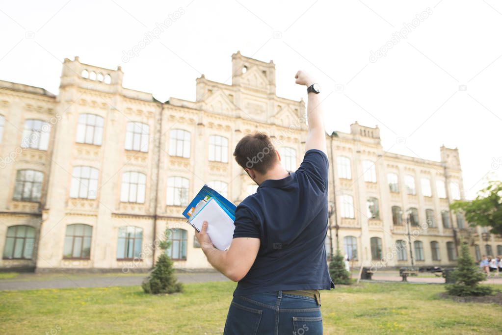 Back of a happy student, with books and notebooks, raised his hands upward with joy against the backdrop of the university building. Joyful man rejoices in the campus of the University